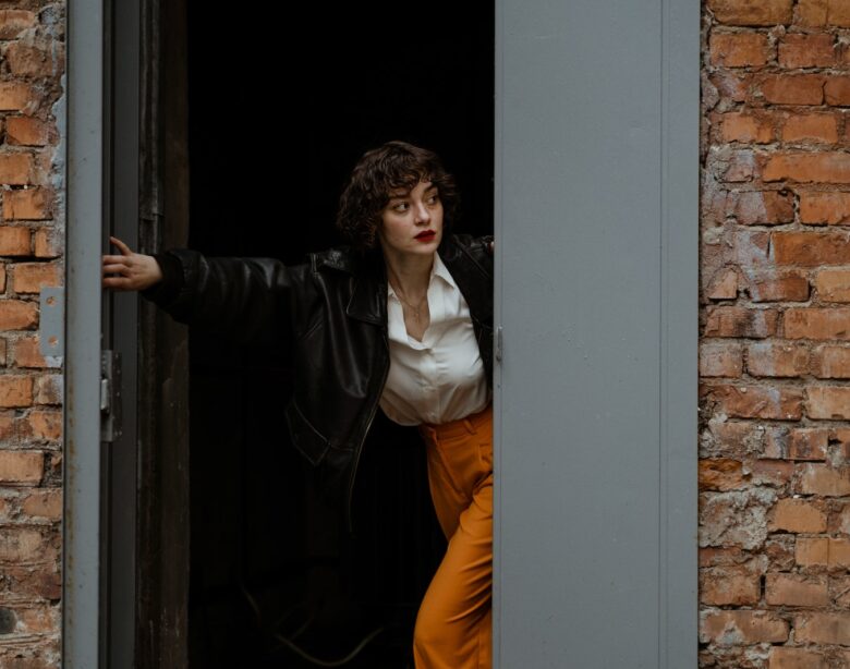 Woman in White Long Sleeve Shirt and Black Leather Jacket Standing on Doorway Looking Outside